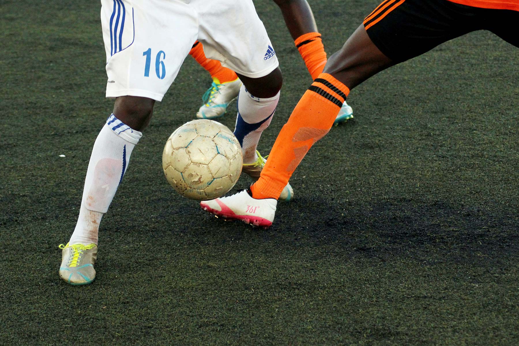 Close-up of two soccer players competing for the ball on a dark playing field. One wears a white uniform with the number 16, the other wears an orange and black uniform.