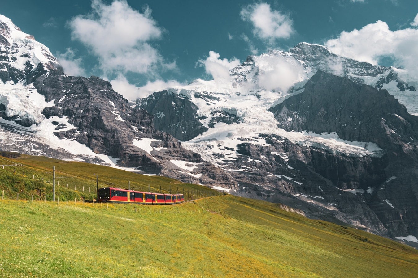 A red and yellow train travels through a lush green alpine meadow beneath dramatic snow-covered peaks in the Swiss Alps, with clouds drifting above the rugged mountain landscape.