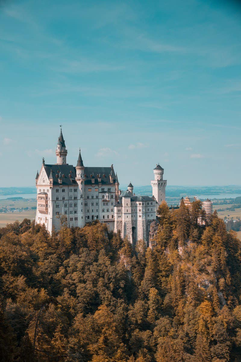 Neuschwanstein Castle perched on a forested hilltop in Bavaria, Germany, with its fairy-tale towers and spires set against a clear blue sky and scenic valley in the background.