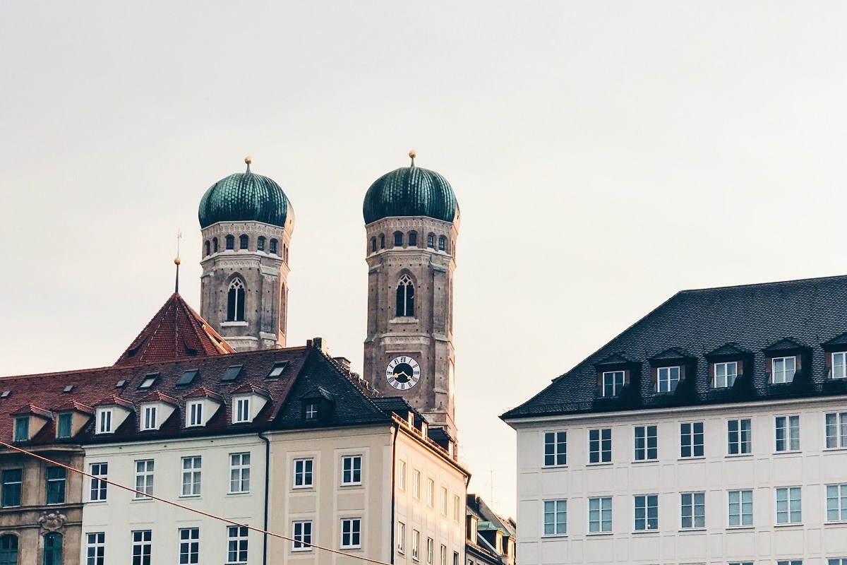 View of the Frauenkirche in Munich, recognizable by its two iconic green domed towers rising behind traditional Bavarian buildings.