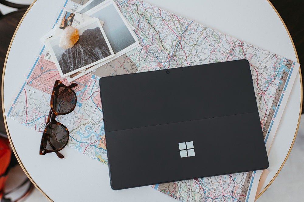A black tablet on top of a folded road map, next to sunglasses and travel photos with a crystal stone on a round white table.