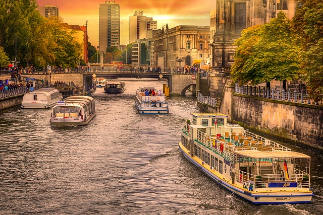 Tour boats cruising along the Spree River in central Berlin at sunset, with historic buildings, trees, and modern high-rises in the background.