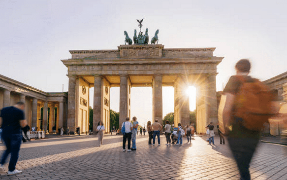 The Brandenburg Gate in Berlin at sunset, with the Quadriga on the roof and Pariser Platz in the foreground. Numerous people stroll or linger around the historic landmark