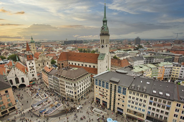 Aerial view of Marienplatz in Munich, Germany, showing the Old Town Hall, St. Peter’s Church with its tall tower, and surrounding historic buildings filled with people and activity under a evening sky.